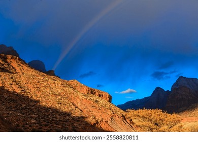 A rainbow is seen in the sky above a rocky mountain range. The sky is mostly blue with some clouds, and the sun is shining through the clouds. The scene is peaceful and serene - Powered by Shutterstock