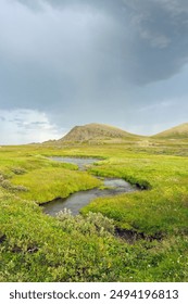 Rainclouds over a hill and a small river flowing trough green landscape at the coast of the Barents Sea in Varanger Peninsula, Northern Norway