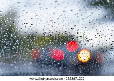 Raindrops cling to a car windshield, distorting the view of bright red and yellow lights from vehicles moving in the rainy city. The atmosphere feels moody and reflective