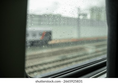 Raindrops cling to a train window while the vehicle passes through a city area, showcasing the blurred scenery outside on a grey day.