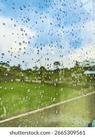 Raindrops cling to a window pane, creating a blurry view of a bright green field and blue sky with white clouds.