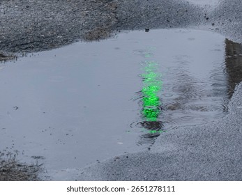 Raindrops create ripples on a puddle reflecting a vibrant green light. The wet urban street has a gray surface and showcases a gloomy yet intriguing atmosphere.