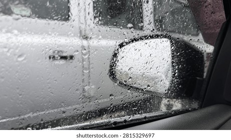 Raindrops dotting a car window, partially obscuring the blurred shape of a white vehicle and its side mirror parked outside.