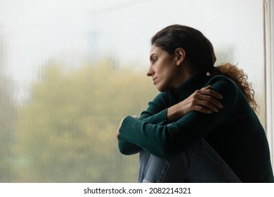 Raindrops like tears. Sad lonely latina female sit on windowsill in melancholic mood watch rain outside feeling her heart broken. Pensive young woman hug herself suffer of unanswered love. Copy space
