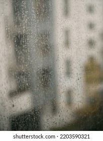 Raindrops On A Window With An Apartment House Background. Concept Of Staying Home While There Is Bad Weather Outside. Cozy Indoor View At Rainy Weather