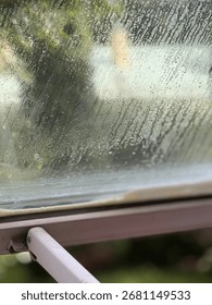 Raindrops on window creating a textured background with blurred green foliage in the distance. Mobile photo