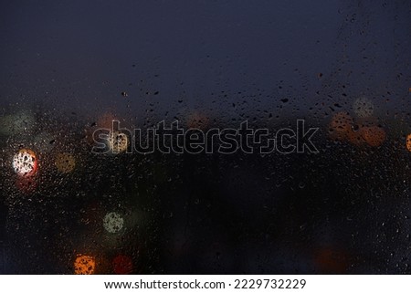 Raindrops on a window glass and unfocused lights of a night or evening city. Blurred Background of night city behind the glass during rain. Rainy weather outside the window