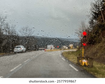 Raindrops on windshield create abstract pattern while driving on wet road with red traffic light and road construction in background