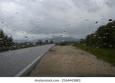 Raindrops on a Windshield Creating a Blurry View of a Countryside Road Under Cloudy Skies on a Rainy Day