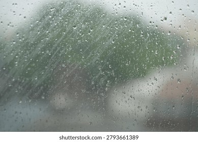 Raindrops resting on a window pane during a rainy day, with a softly blurred outdoor background visible through the glass. The scene captures a calm, peaceful atmosphere and the cozy feeling of staying indoors while watching the rain outside