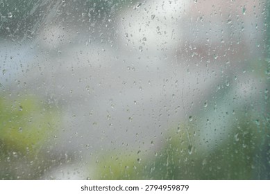 Raindrops resting on a window pane during a rainy day, with a softly blurred outdoor background visible through the glass. The scene captures a calm, peaceful atmosphere and the cozy feeling of staying indoors while watching the rain outside