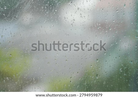 Raindrops resting on a window pane during a rainy day, with a softly blurred outdoor background visible through the glass. The scene captures a calm, peaceful atmosphere and the cozy feeling of staying indoors while watching the rain outside
