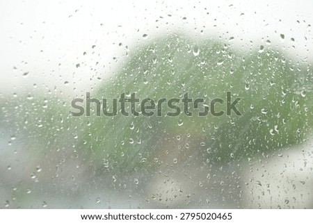 Raindrops resting on a window pane during a rainy day, with a softly blurred outdoor background visible through the glass. The scene captures a calm, peaceful atmosphere and the cozy feeling of staying indoors while watching the rain outside