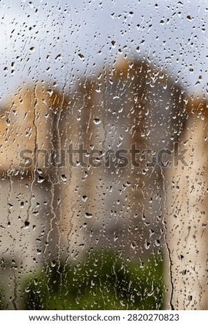Raindrops streak down a windowpane, blurring the view outside. Faint outlines of buildings and greenery appear through the wet glass. The scene feels quiet and introspective