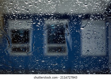 It's Raining Outside, Through The Wet Glass You Can See A House With Three Windows, One Of Them Is Closed With Metal Blinds, Selective Focus On Raindrops, Urban Landscape