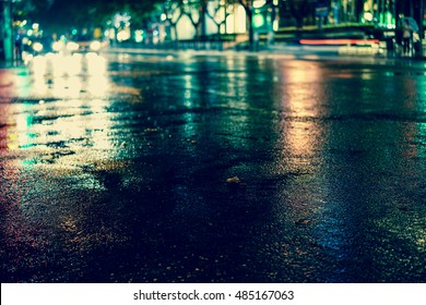Rainstorm in the big city night, light from the shop windows reflected on the road on which cars travel. View from the level of asphalt