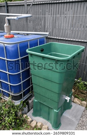 Rainwater overflow system connecting two water storage tanks. The green tank collects excess from the blue container for sustainable garden use.