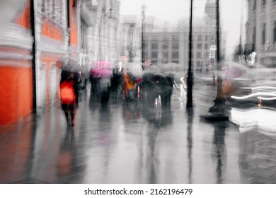 Rainy Cold Evening In The City. Unrecognizable Silhouettes Of People Under Umbrellas Walking Along The Street, City Life. Abstract Blurred Background. Intentional Motion Blur