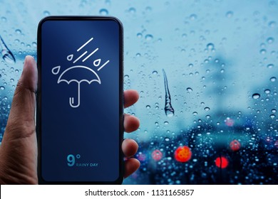 Rainy Day Concept. Hand Holding Smartphone with Weather Information show on Screen. Blurred Traffic Jam and Rain Drops on Glass Window as background 
