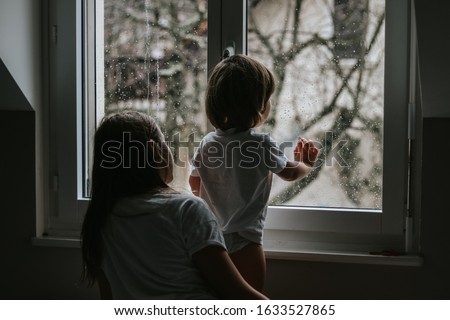 Rainy day. Little toddler boy with his sister looking outside through  wet window from his room missing mother.  Siblings love, care and support. Lifestyle. Quarantine and isolation period at home