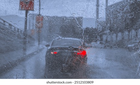 A rainy day street scene captured from inside a car, with raindrops on the window blurring the view of a car with red brake lights on a wet road. The image conveys a sense of calm and the typical 