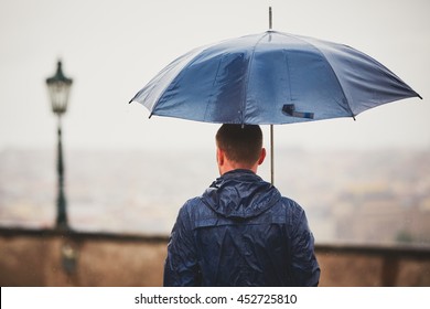 Rainy Day. Young Man Is Holding Blue Umbrella And Walking In Rain. Street Of Prague, Czech Republic. - Selective Focus On Person