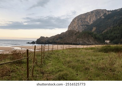 A rainy evening at Laga Beach in Basque Country, Spain. Moody clouds with a soft pink hue cover the sky. The foreground shows lush greenery, leading to the beach, rocky shoreline and distant mountains - Powered by Shutterstock