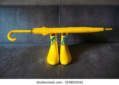 Rainy Mood, outfit for rainy day. Blue raincoat, yellow rubber boots and umbrella on a gray sofa. Colors of the year 2021 Ultimate Gray and Illuminating background.