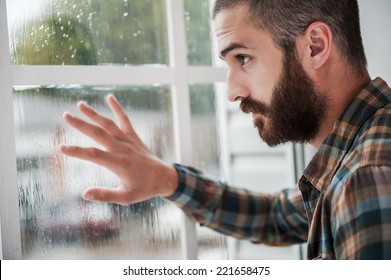 Rainy mood. Thoughtful young bearded man touching the window and looking through it - Powered by Shutterstock