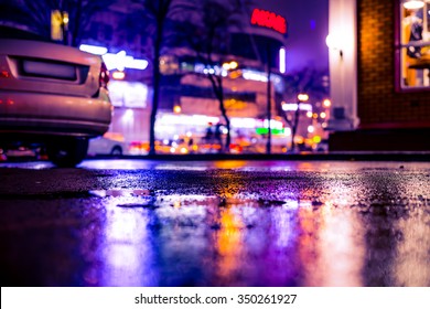 Rainy Night In The Big City, City Alley With Trees And A Parked Car Near The Loaded Avenue With Shops. View From The Level Of Asphalt, Image In The Soft Orange-purple Toning