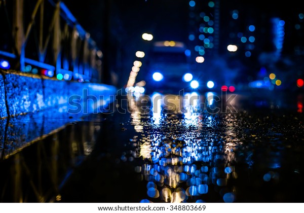 Rainy night in the big city, approaching\
headlights of cars traveling along the avenue. View from the level\
of the curb on the road, in blue\
tones