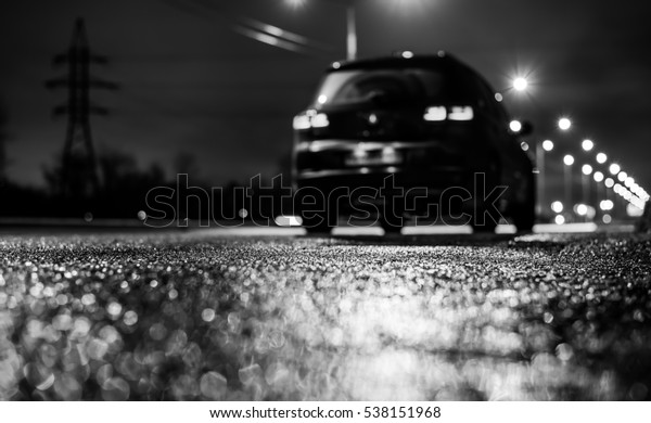 Rainy night in the big city, the big black car\
parked on the roadside. Close up view from the asphalt level, image\
in the black and white\
tones