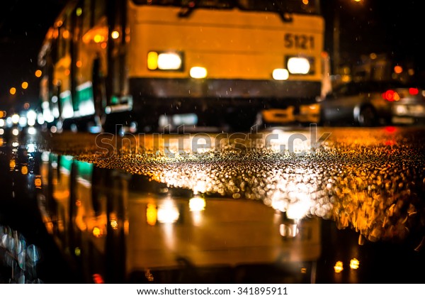 Rainy night in the big city, city\
bus comes to a halt. View from puddles on the pavement\
level