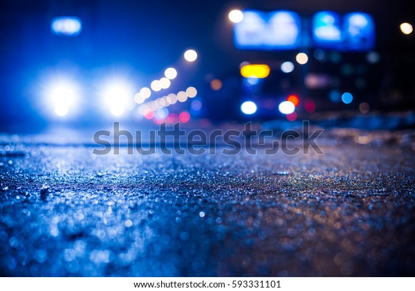 Rainy night in\
the big city, glare from the headlights of the parked car and a\
passing near the stream of cars. Close up view from the sidewalk\
level, image in the blue\
tones