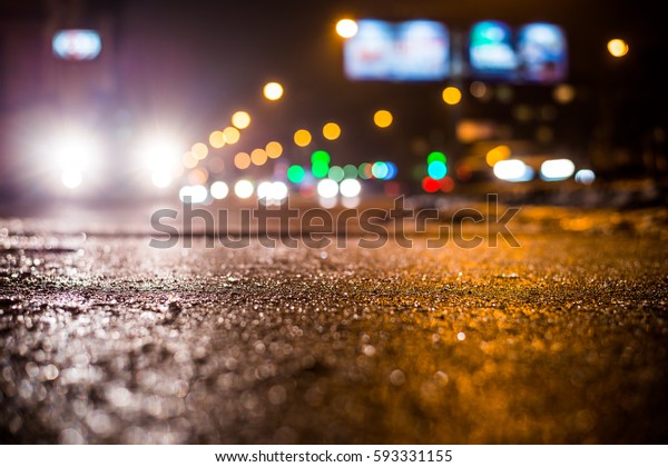 Rainy night in the big city, glare from\
the headlights of the parked car and a passing near the stream of\
cars. Close up view from the sidewalk\
level