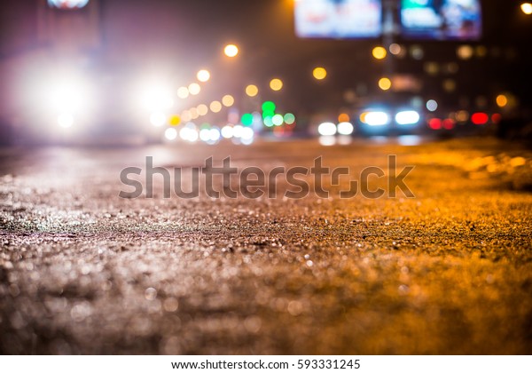 Rainy night in the big city, glare from\
the headlights of the parked car and a passing near the stream of\
cars. Close up view from the sidewalk\
level