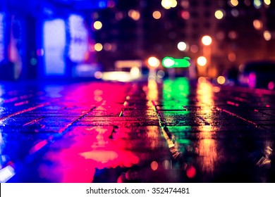 Rainy Night In The Big City, Light From The Night Club And The Windows Of The House Is Reflected In The Asphalt. View From The Sidewalk Level Paved With Bricks, Image In The Yellow-blue Toning