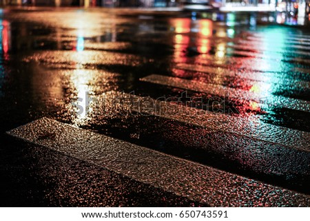 Rainy night in the big city, light from the shop windows reflected on the road on which cars travel. View from the level of asphalt.