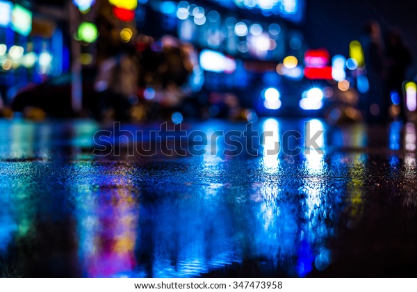 Rainy night in the big city, light from the shop\
windows reflected on the road on which cars travel. View from the\
level of asphalt, in blue\
tones