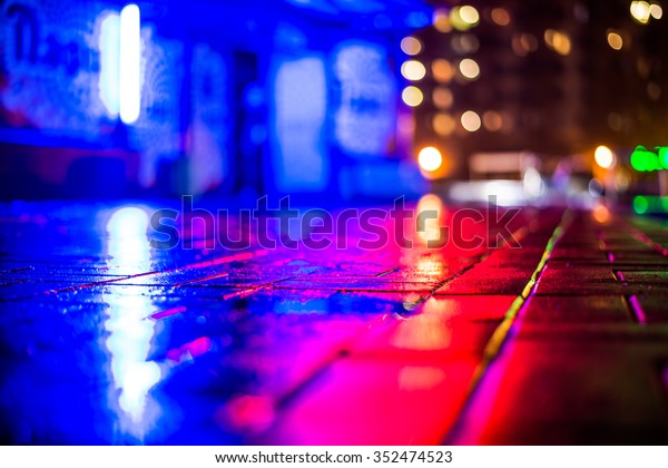 Rainy night in the big city, light from\
the night club and the windows of the house is reflected in the\
asphalt. View from the sidewalk level paved with\
bricks