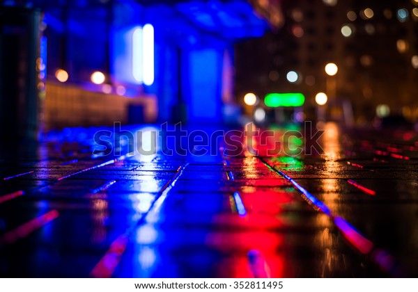 Rainy night in the big city, light from\
the night club and the windows of the house is reflected in the\
asphalt. View from the sidewalk level paved with\
bricks
