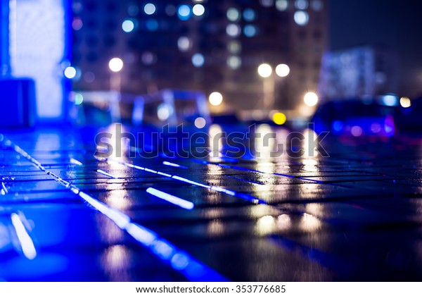 Rainy night in the big\
city, light from the night club and the windows of the house is\
reflected in the asphalt. View from the sidewalk level paved with\
bricks, in blue tones