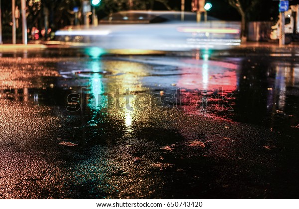 Rainy night in the big city, light from the shop\
windows reflected on the road on which cars travel. View from the\
level of asphalt.