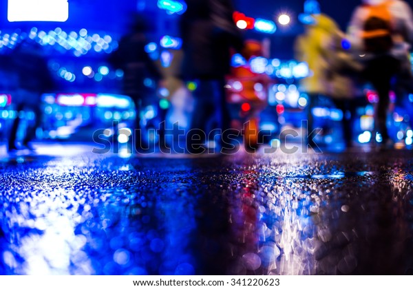 Rainy night in the big city, pedestrians cross the\
busy intersection in the light of shop windows. View from the level\
of asphalt, in blue tones