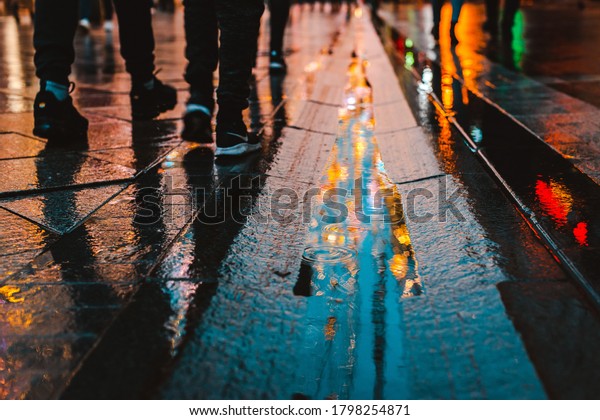 Rainy night in a big city, reflections of\
lights on the wet road surface. The view from the street level.\
Pedestrian feet and abstract\
background.