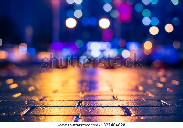 Rainy night in the\
city. Light from the windows of the house opposite. Car parking.\
Multi-colored shop windows. Close up view from the pavement level.\
Orange-blue toning.