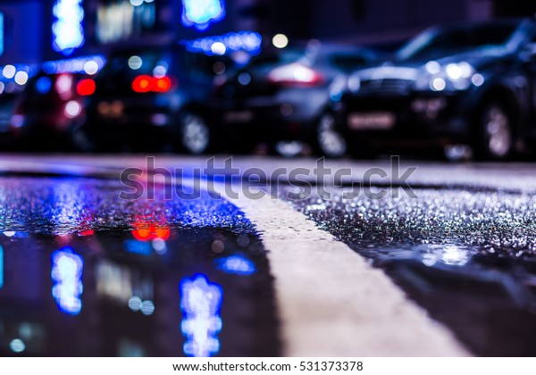 Rainy night in the\
parking shopping mall, parked cars illuminated advertising signs.\
Close up view of a puddle on the level of the dividing line, image\
in the blue tones