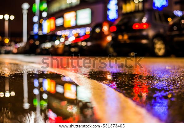 Rainy night in the parking\
shopping mall, parked cars and marking the line going into the\
distance. Close up view of a puddle on the level of the dividing\
line