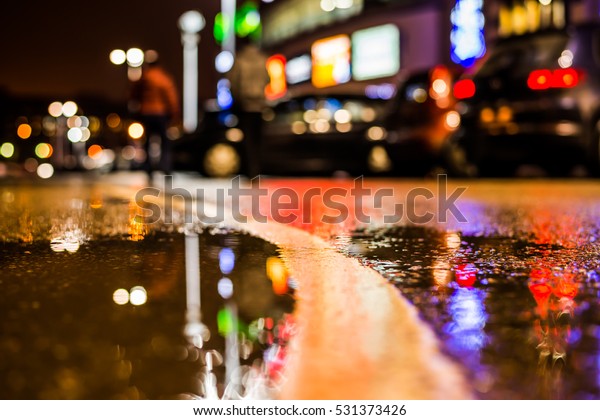 Rainy night in the parking shopping mall, parked\
cars and people walking. Close up view of a puddle on the level of\
the dividing line