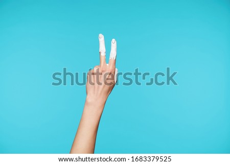 Raised female's elegant hand forming figure two with fingers in body language, having white paint on raised fingers while posing over blue background